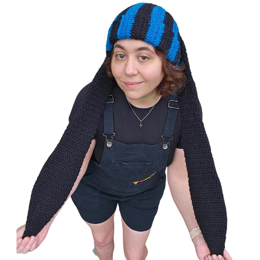 The image features a crocheted beanie with extra long bunny ears in a black and sapphire blue striped pattern. The ears are stretched to their max length.